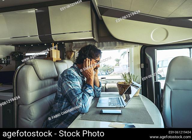 Adult man sitting at the desk using laptop computer and mobile phone connection to work in alternative office inside a camper van