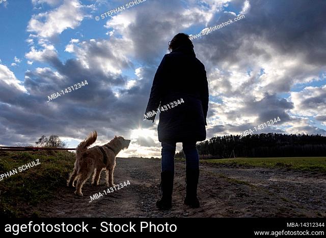 A woman stands with her dog on a country lane, while on the horizon the evening sun breaks through the clouds