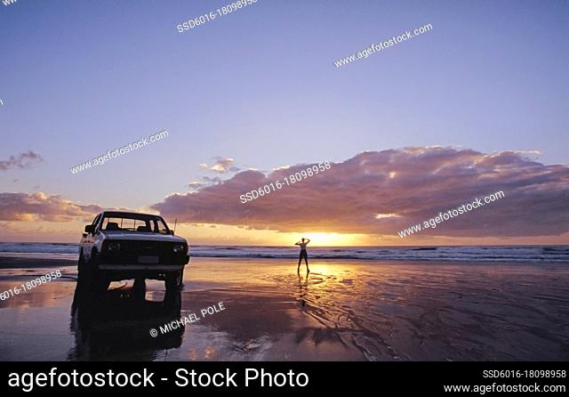 White Ute parked on the beach and lone person standing by the water at sunset