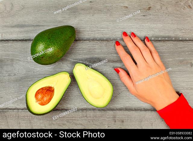 Table top view - avocado cut in half, woman hand with red nails next to it, placed on gray wooden desk