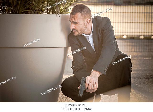 Man in business attire behind plant pot holding handgun