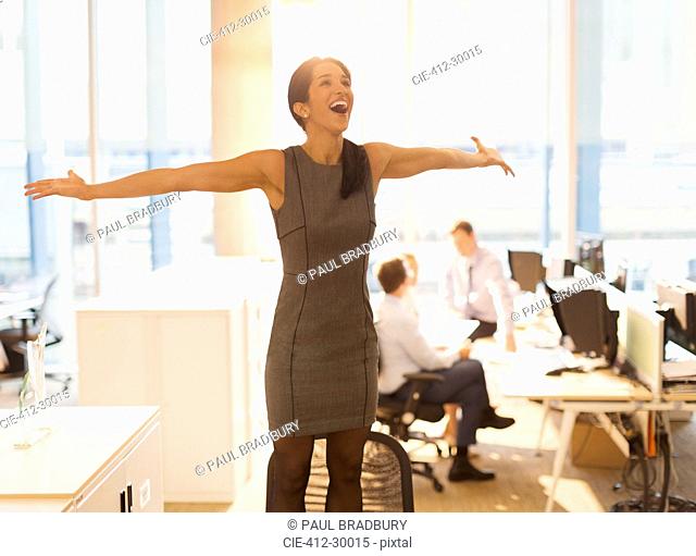 Exuberant businesswoman celebrating with arms outstretched on top of chair in office