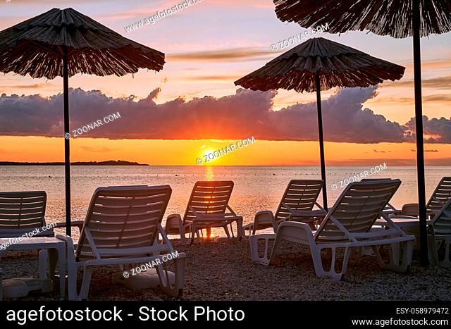Straw beach umbrellas and chairs on a sunset beach with beautiful violet clouds