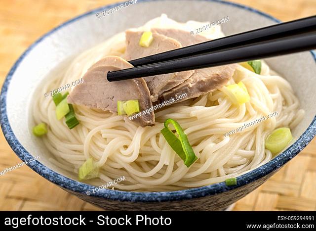 A studio photo of chop sticks grabbing a pork slice on top of a bowl of noodles