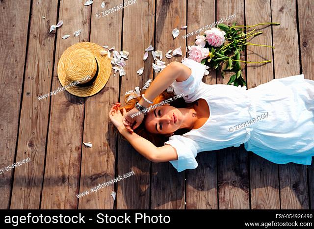 Attractive young hipster woman in white dress and straw hat lies on the wooden floor