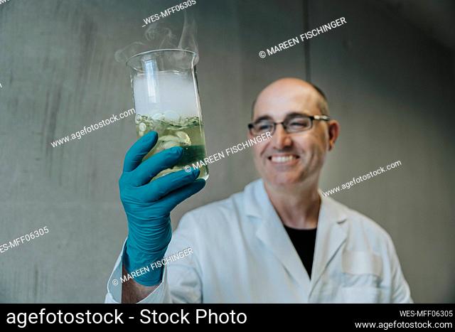 Smiling scientist holding liquid flask with evaporating smoke while standing at clinic corridor