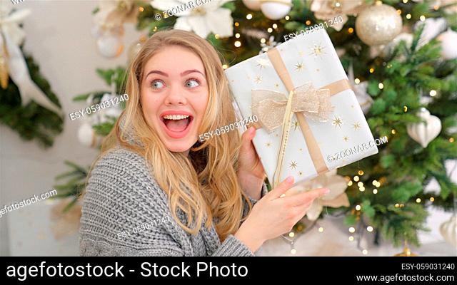 Surprised and happy young woman with christmas present box near christmas tree. Xmas home concept