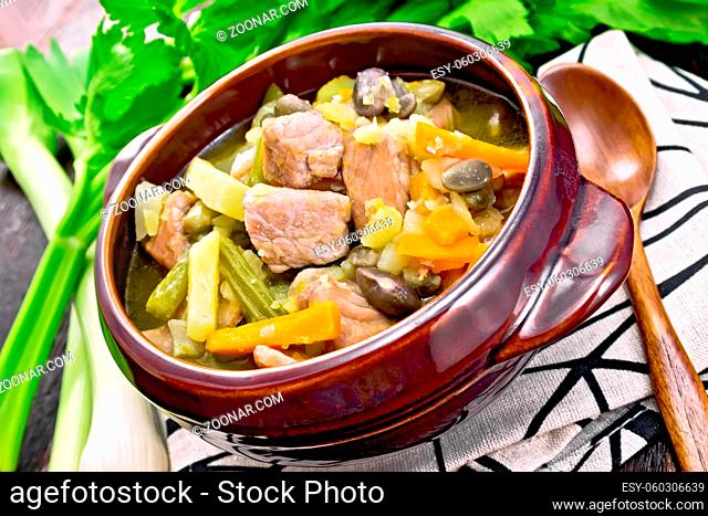 Eintopf soup of pork, celery, beans, carrots and potatoes with leek in a clay bowl on a towel on background of dark wooden board