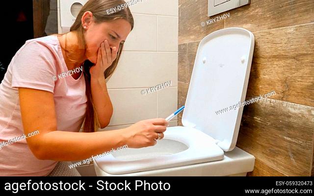 Young woman looking on positive pregnancy test sitting on toilet and holding her sickness. Intoxication and nausea during pregnancy