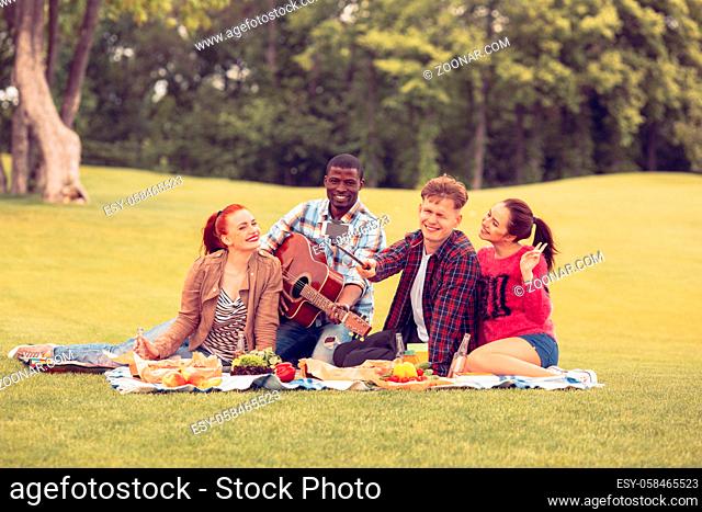 Picture of happy best friends sitting on picnic rug and smiling for camera. People resting and relaxing on picnic with fruits, vegetables and alcoholic drinks