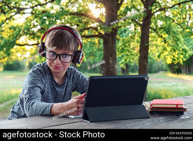 Surprised junior pupil in glasses and earphones looking at screen of tablet while sitting at park table. Boy with dark hair wearing gray longsleeve amazed by...