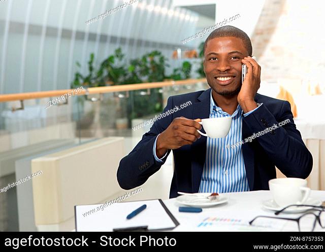 Businessman speaking over mobile phone while drinking hot cup of coffee. Happy handsome man in business suit looking at camera in restaurant or cafe