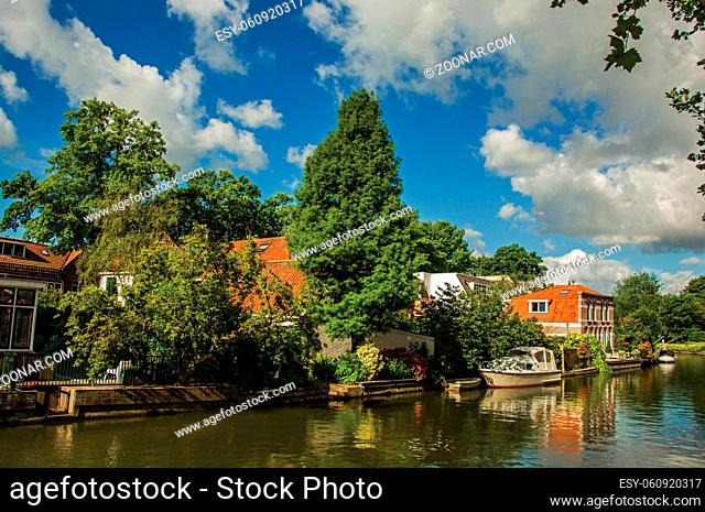 Canal with trees, brick houses and boat moored on its bank reflected in water and blue sky of sunset in Weesp. Quiet and pleasant village full of canals and...