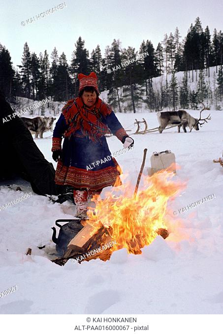 Finland, Saami woman standing next to fire, sled reindeer in background