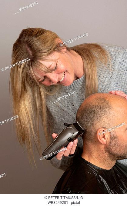 Woman using portable electric hair clippers to give a balding man a crew cut