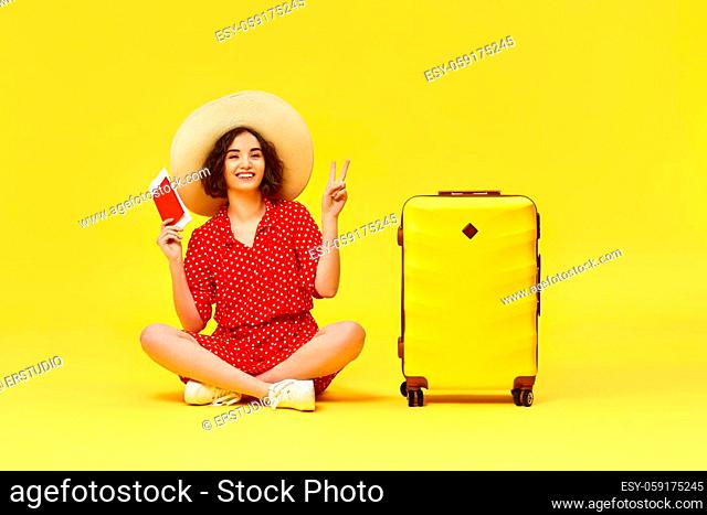 excited woman in red dress with suitcase and passport sitting on floor on yellow background. concept of travel