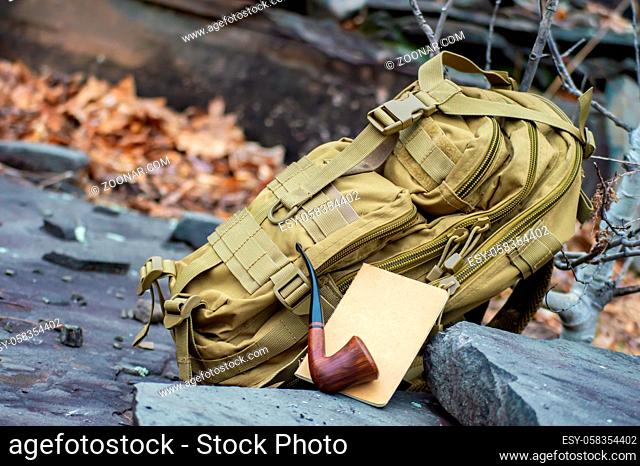 Close up still life style shot of a green army tactical backpack, a travel notebook and a wooden vintage pipe disposed on rock steps outdoor