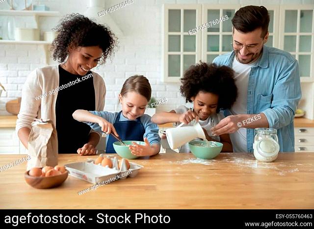 Happy young multiracial parents and little biracial daughters have fun baking in kitchen together, overjoyed family with cute small multiethnic children cooking...