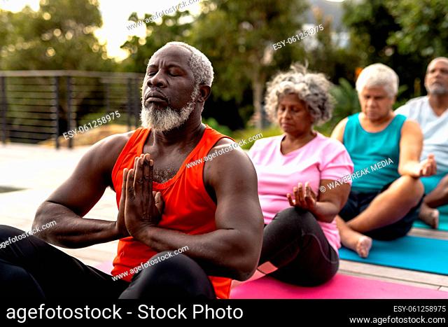 Senior multiracial friends meditating while sitting on mats against plants in yard at nursing home