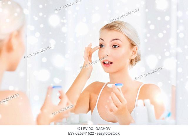 beauty, vision, eyesight, ophthalmology and people concept - young woman putting on contact lenses at mirror in home bathroom over snow