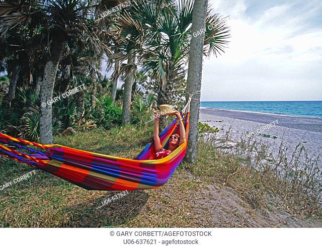 Woman in a hammock in Florida at the beach holding up a straw hat and looking