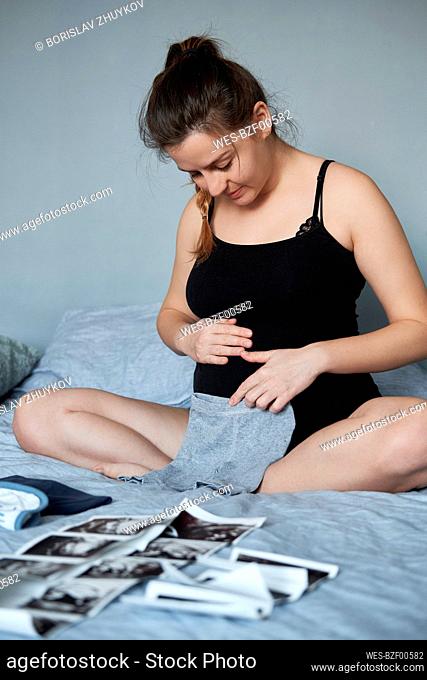 Pregnant woman holding baby clothes on stomach while sitting with ultrasound scans on bed at home