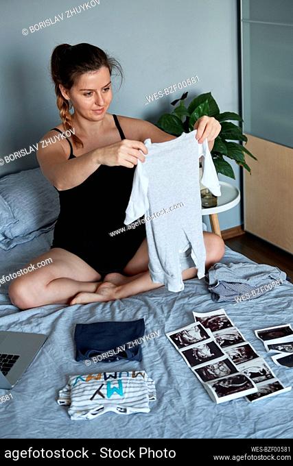 Pregnant woman preparing clothes for her baby boy on bed at home