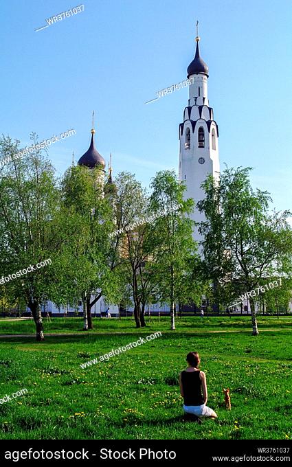 a young woman with a small dog sitting on grass in the park with a church in the background