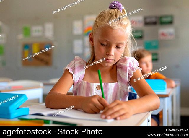Caucasian elementary schoolgirl writing on book at desk in classroom