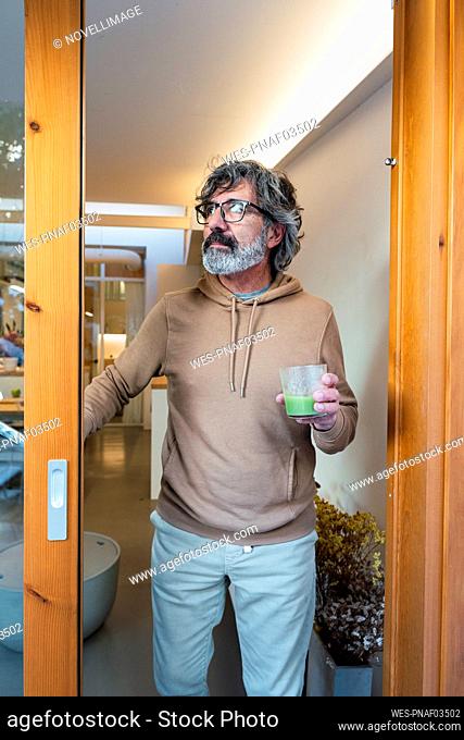 Man with glass of smoothie standing by sliding door at home
