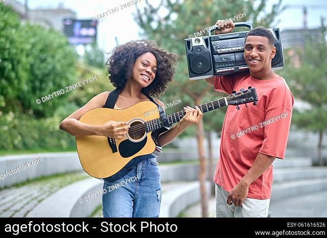 Happy people. Young dark-skinned girl playing guitar and guy with record-player on shoulder smiling at camera standing in park