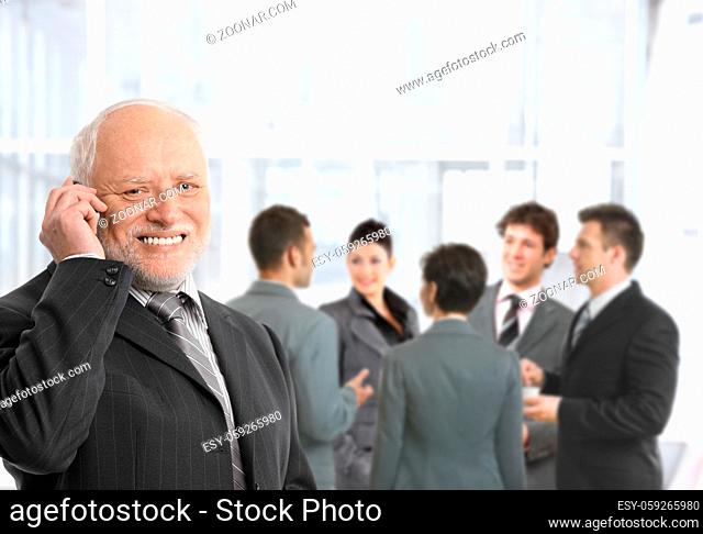 Senior businessman talking on mobile phone in office lobby, smiling, businesspeople chatting in background