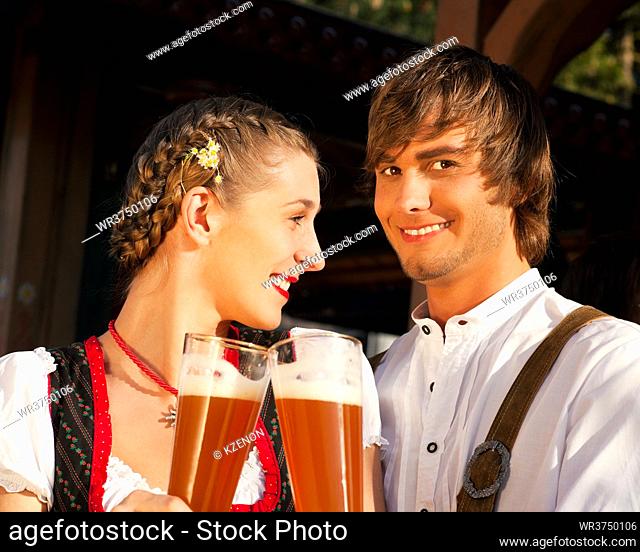 Couple in traditional Bavarian Tracht - Dirndl and Lederhosen - in front of a beer tent at the Oktoberfest or in a beer garden enjoying a glass of tasty wheat...