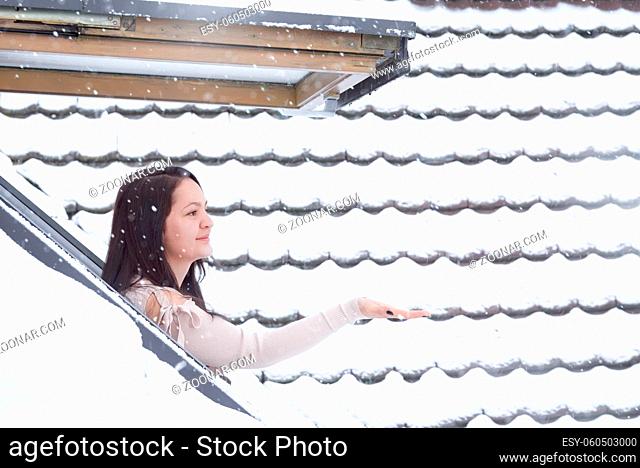 Attractive brunette woman dressed in a cozy sweater standing outside on her window reaching her hand to catch the snowflakes