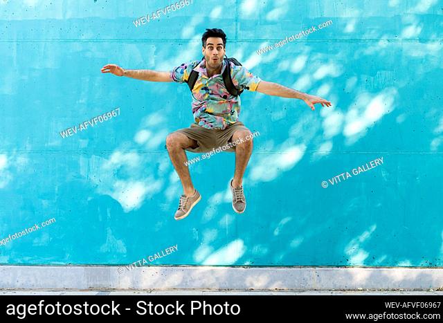 Happy man with backpack jumping in front of blue wall
