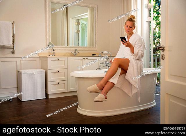 Caucasian woman sitting in bathroom wearing bathrobe using smartphone, running a bath