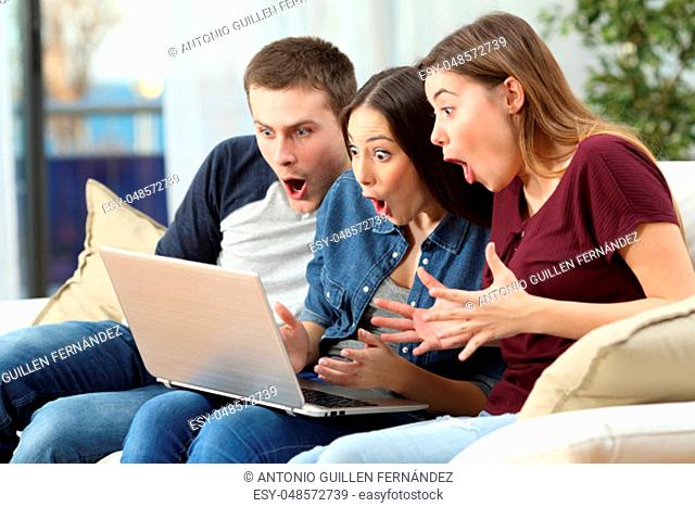 Three amazed friends watching media content on line in a computer sitting on a couch in the living room at home