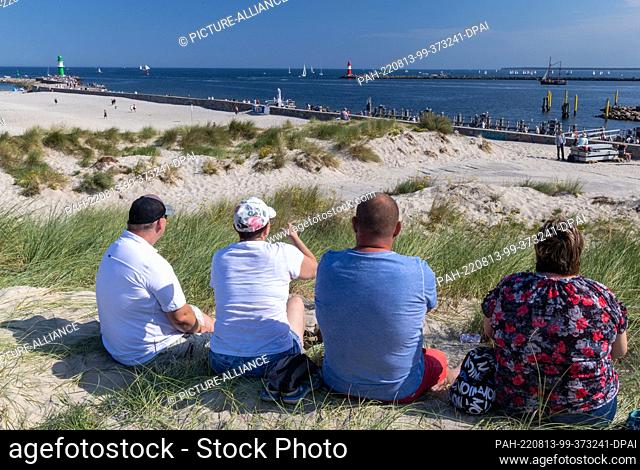 13 August 2022, Mecklenburg-Western Pomerania, Rostock: Visitors sit in the dunes and watch the sailing ships leaving for guest cruises on the Baltic Sea within...