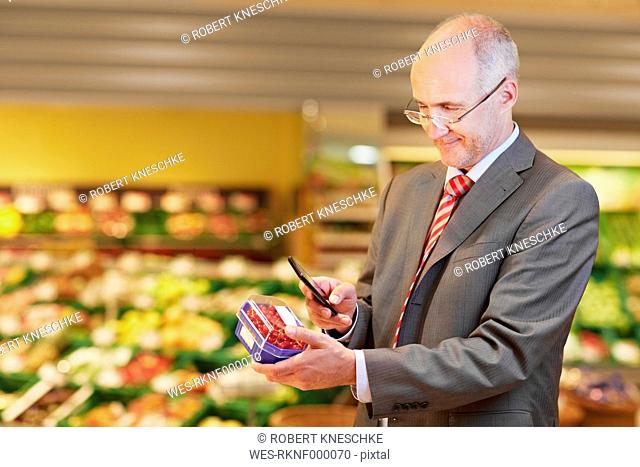 Germany, Cologne, Mature man with redcurrant and mobile in supermarket