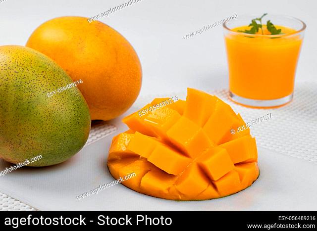 Photo of half a mango, two mangoes behind and a glass of mango juice in the background