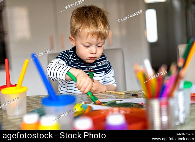 Three year old boy busy painting at home, with paint pots and brushes