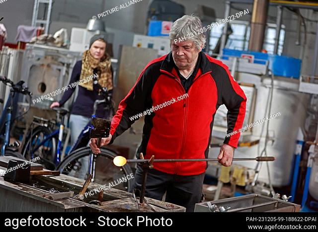 05 December 2023, Saxony-Anhalt, Derenburg: A glassmaker works at the furnace and shapes a piece of glass while a visitor on a bicycle watches him