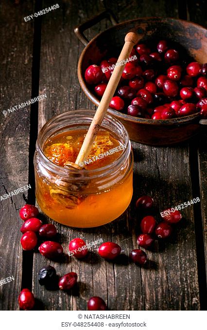 Open glass jar of liquid honey with honeycomb and honey dipper inside and fresh cranberries in vintage bowl over old wooden table. Dark rustic style