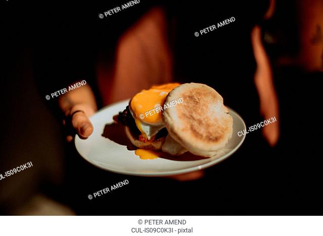 Barista holding plate with fried egg muffin in cafe, close up low key