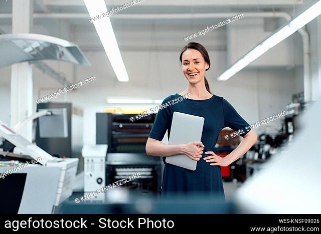 Happy businesswoman holding digital tablet while standing with hand on hip in industry