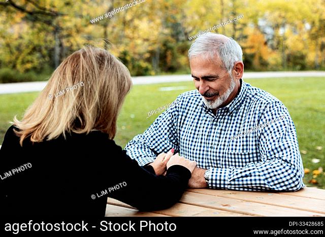 A mature couple praying together at a picnic table on a warm fall day in a city park; St. Albert, Alberta, Canada