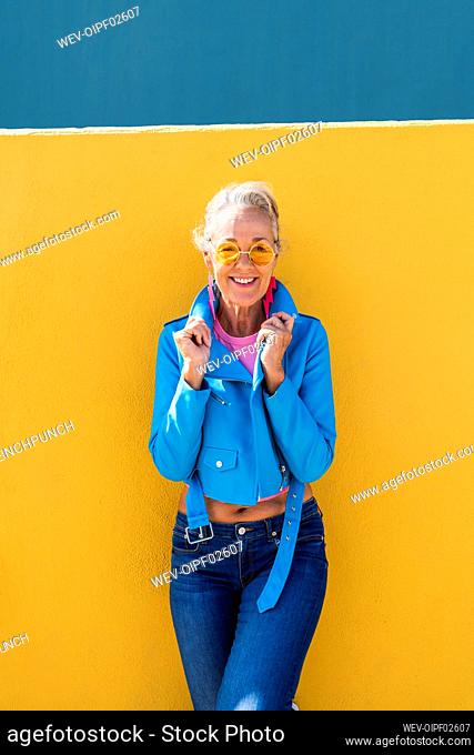 Smiling mature woman wearing blue leather jacket leaning on colored wall