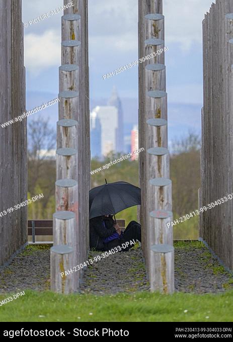 PRODUCTION - 13 April 2023, Hesse, Dreieich: Two people sit under an umbrella between the tree trunks of the rod pyramid and look at the skyline of Frankfurt am...