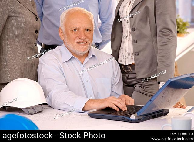 Portrait of senior businessman working on laptop computer, looking at camera, smiling, coworkers standing behind