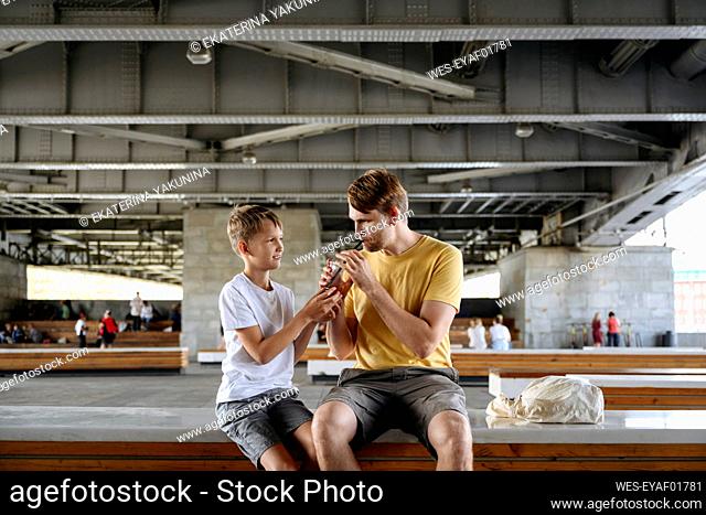 Smiling boy feeding iced coffee to father while sitting on bench